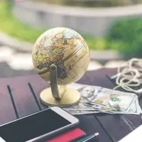 An image of a globe, cell phone, headphones, and money sitting on a desk providing an overall travel theme.