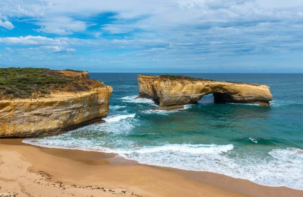 Two rock formations in the water with golden sand, known as London Bridge, as one of many Great Ocean Road stops.