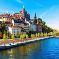 Featured image for this Stockholm bucket list - The buildings of Stockholm on the left side of the image and a sidewalk and river on the right hand side of the image.