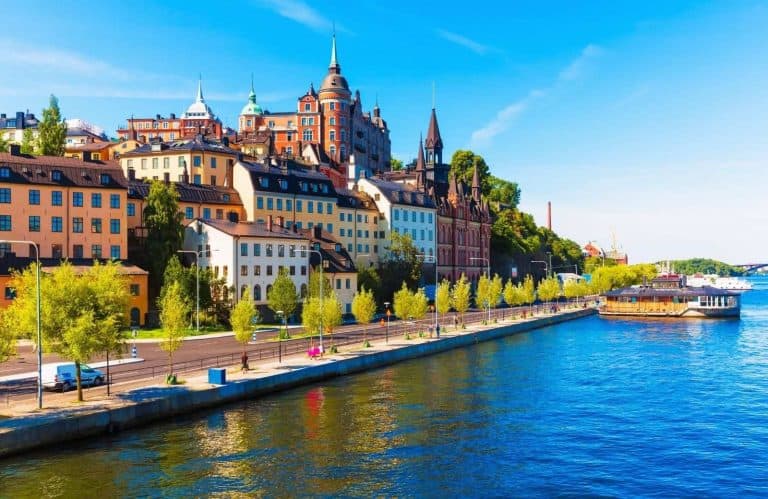 Featured image for this Stockholm bucket list - The buildings of Stockholm on the left side of the image and a sidewalk and river on the right hand side of the image.
