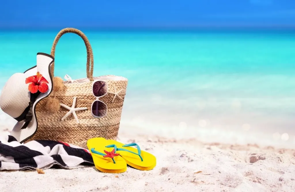 A brown beach bag, yellow flip flops, a beach hat on top of the sand in front of turquoise waters.