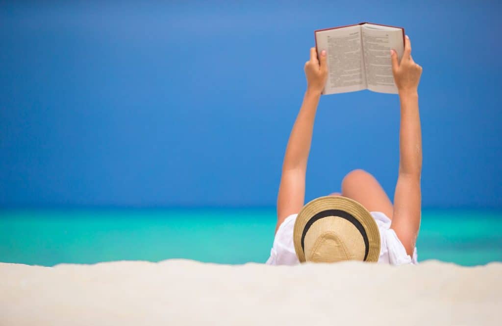 A woman wearing a beach hat laying on the beach reading a book which belongs on a Maldives packing list.