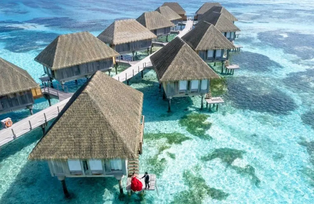 An aerial view of overwater villas in the Maldives with a woman in a red dress and a man in a suit.