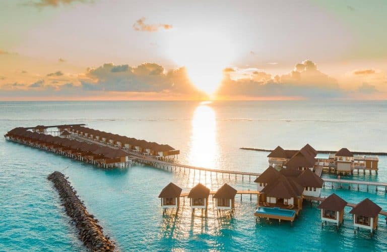 Overwater bungalows with brown thatched roofs on the Indian Ocean in the Maldives with the sun behind them.