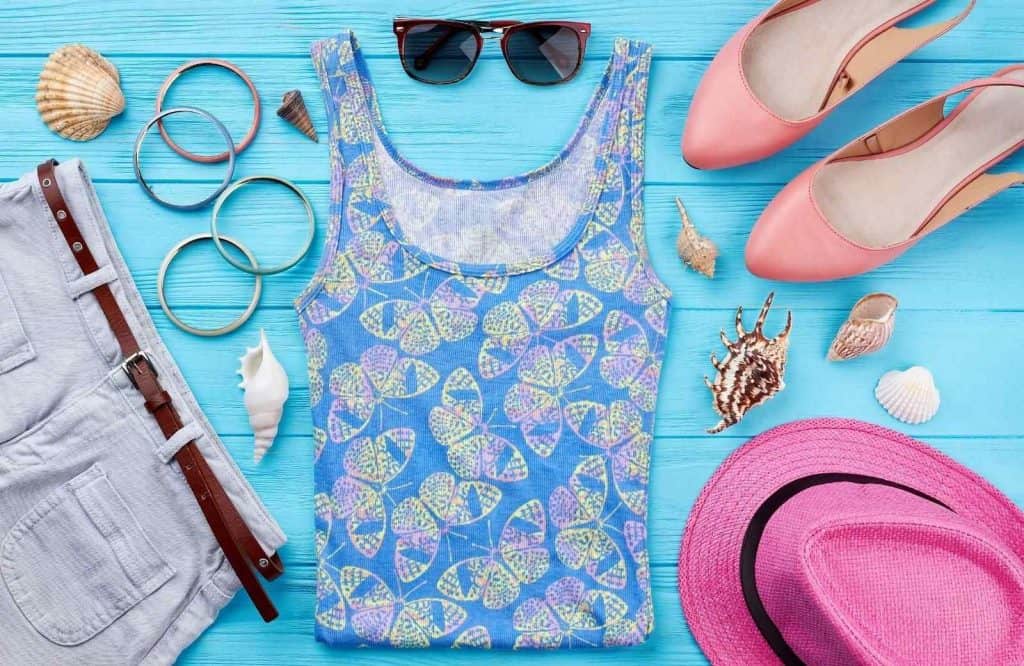 A pair of jean shorts, a blue patterned tank top, a pink hat, and pink shoes against a blue background indicate things to add to your Maldives packing list.