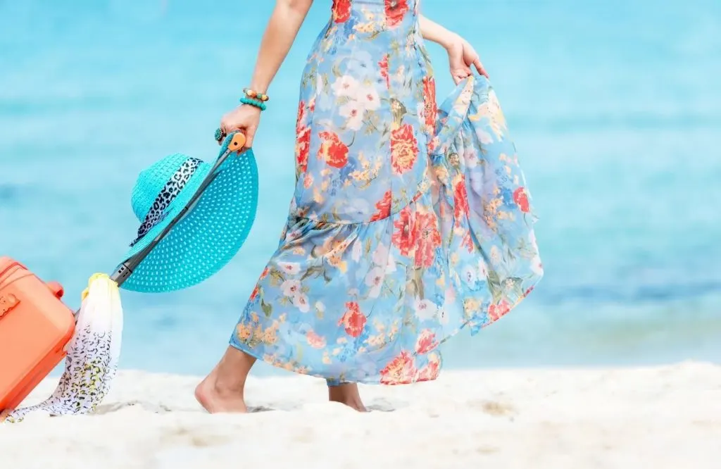 A woman wearing a floral dress dragging a suitcase on the beach for solo travel quotes