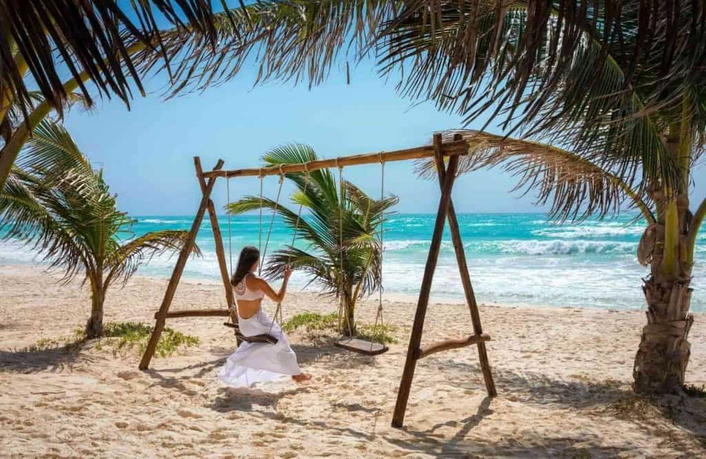 A woman in a white dress on a swing on the beach for a Tulum packing list.