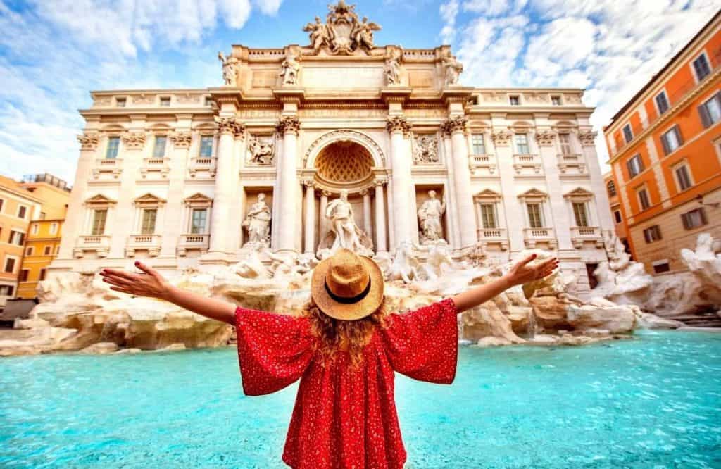 The Trevi Fountain with a blue water fountain and white building with a girl in a red shirt with her arms out, what is Italy famous for.