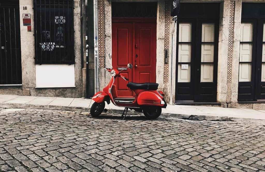 A red Vespa parked on a cobblestone street in front of a red door showing what is Italy famous for.