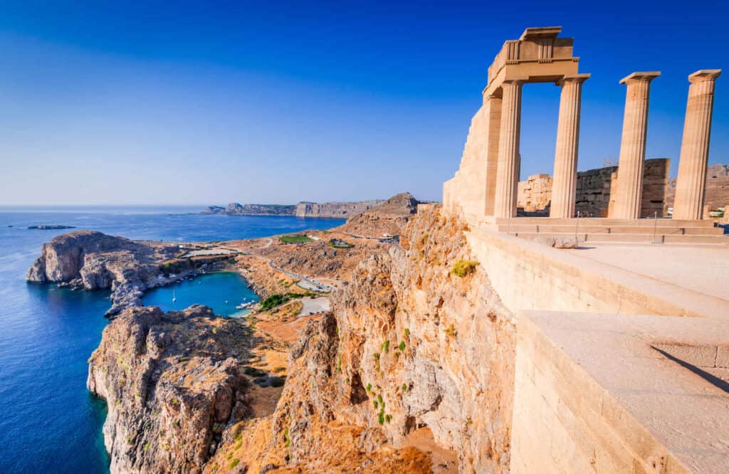 The ancient ruins of Lindos Acropolis in Rhodes with four tall columns on a cliff overlooking the sea.