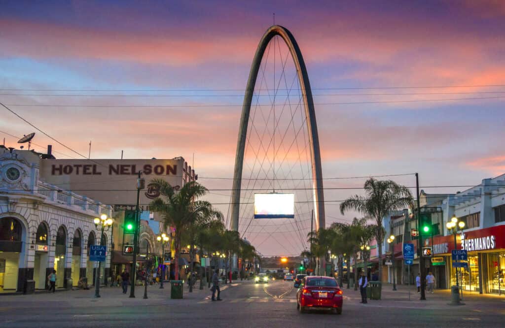 A big arch in the middle of Avenida Revolucion in Tijuana at sunset with shops and businesses on each side of the street.