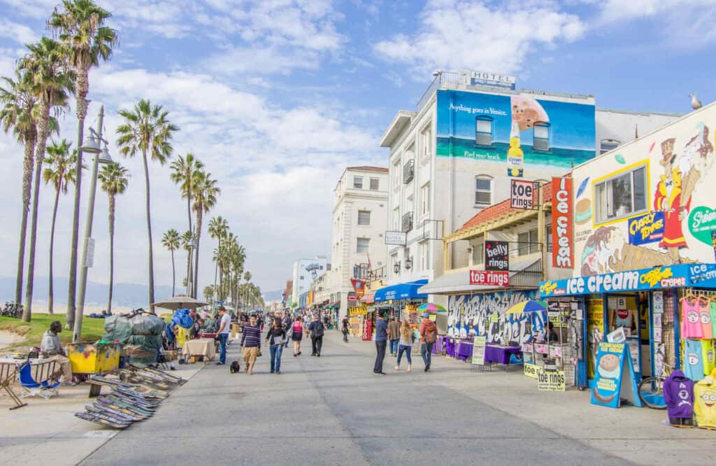 A boardwalk with shops on one side and palm trees on another which is the Ocean Front Walk in Venice Beach.