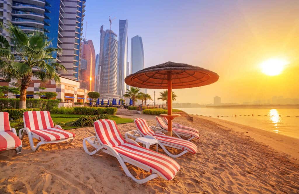 Sunrise on a beach in Abu Dhabi with towering skyscrapers and a red and white beach chairs under a red umbrella.