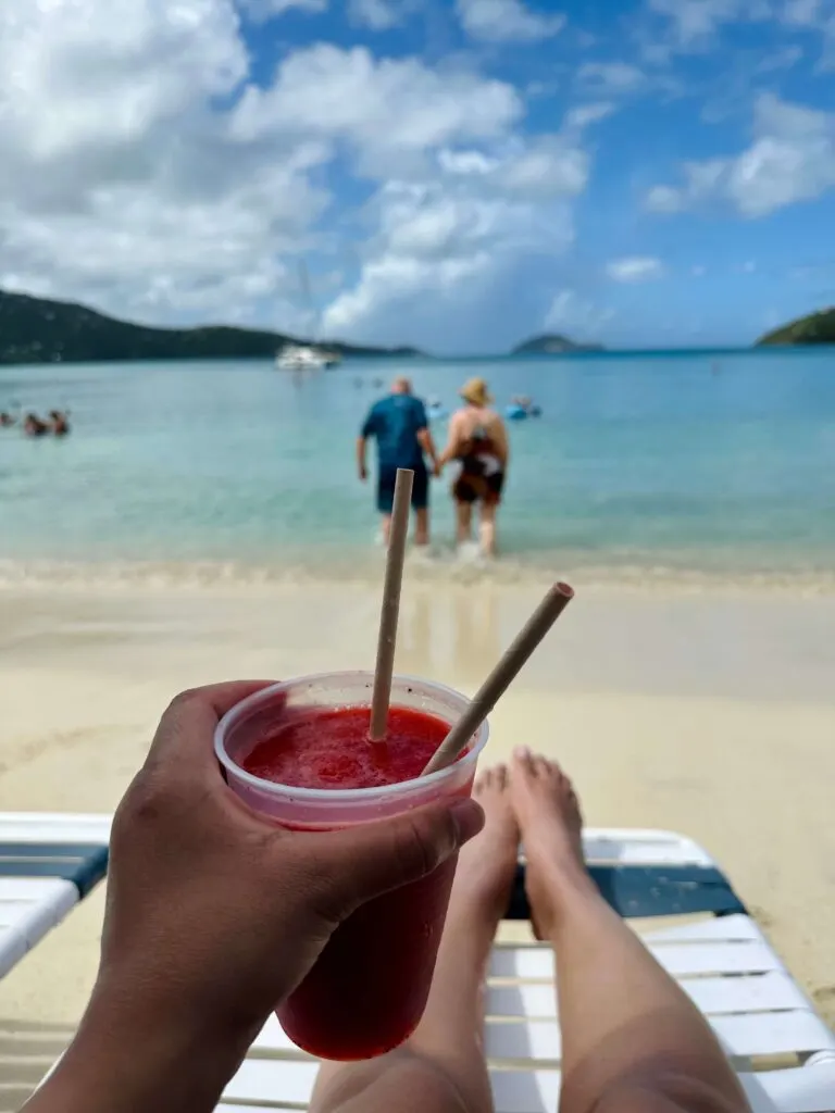 A woman sitting on a beach chair at Magens Bay Beach holding a strawberry daiquiri with the beach in the background, which is one of many things to do on St Thomas Virgin Islands.