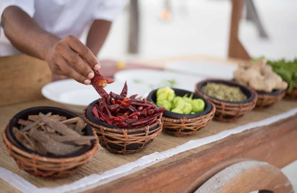 A cooking class where the participant is holding dried red chili peppers with a bowl of four other spices.