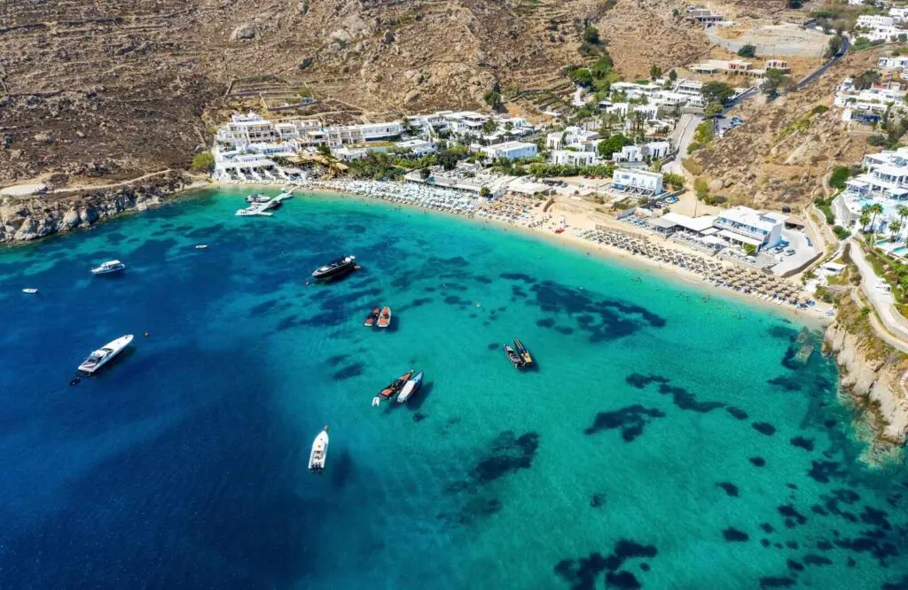 Aerial view of a pristine turquoise beach in Mykonos with boats and a golden shoreline.