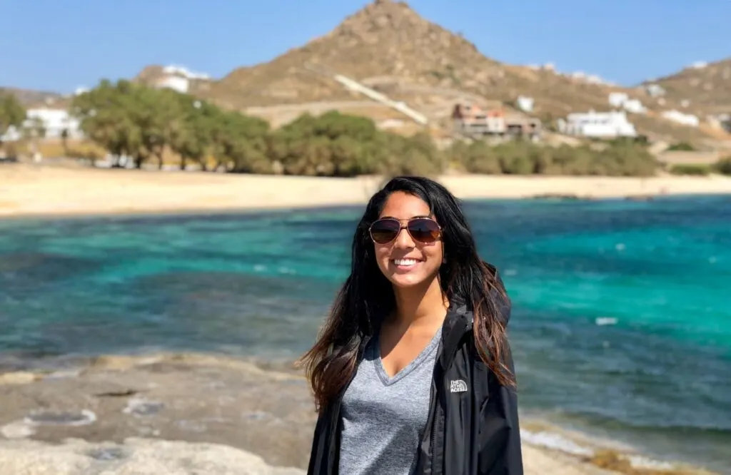 A woman standing in front of a beach with a mountain in the background, which is Kalafatis Beach in Mykonos.