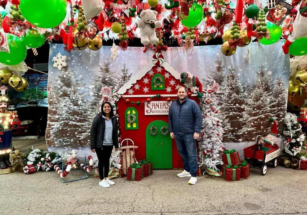 A woman and man standing on either side of a wooden cutout that looks like Santa's workshop under giant green and red ornaments at the Santa Chute.