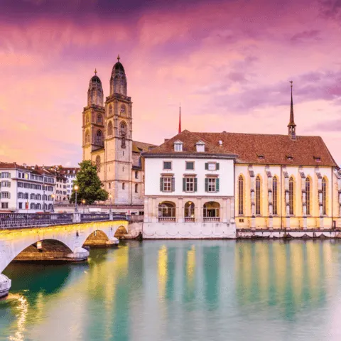 The image depicts a scenic view of Zurich, Switzerland, showcasing the iconic Grossmünster church with its twin towers, a bridge over the Limmat River, and traditional buildings along the water. The sky is painted with vibrant hues of pink and purple, reflecting the setting sun. The calm water and historical architecture create a picturesque, serene atmosphere.