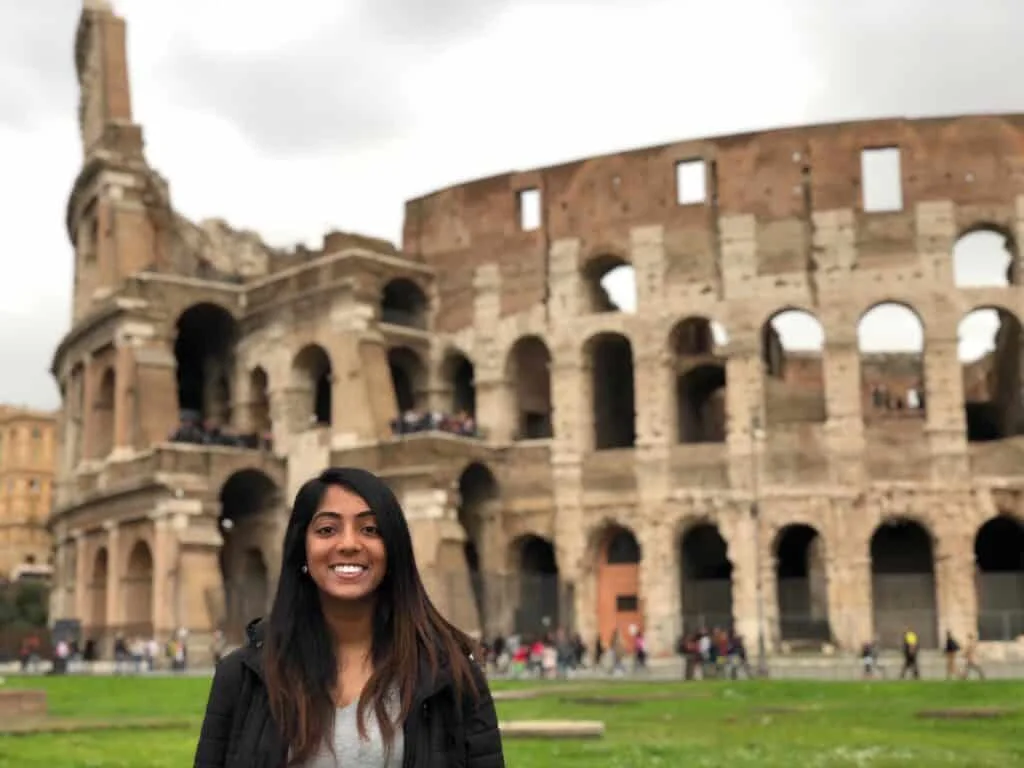 A traveler smiles in front of the iconic Colosseum in Rome, a must-see stop on any one day in Rome itinerary, with the ancient structure's arches and crowds of tourists highlighting its historic charm.