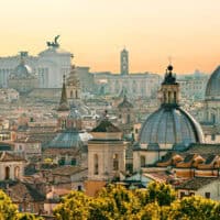 A panoramic view of Rome’s historic cityscape at sunset, featuring a cluster of domed churches and classical buildings with terracotta rooftops. The white marble Altare della Patria, with its iconic winged chariots, stands prominently in the background, bathed in warm golden light.