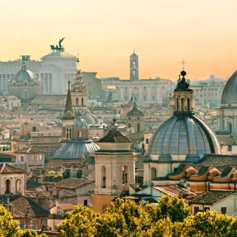 A panoramic view of Rome’s historic cityscape at sunset, featuring a cluster of domed churches and classical buildings with terracotta rooftops. The white marble Altare della Patria, with its iconic winged chariots, stands prominently in the background, bathed in warm golden light.