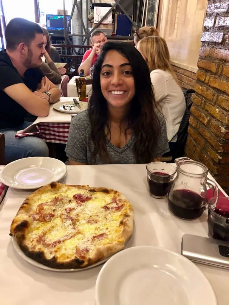 Smiling woman seated at a cozy Roman trattoria with a wood-fired pizza topped with cheese and prosciutto, and a carafe of red wine on the table, capturing a classic meal during one day in Rome. The restaurant has a warm, rustic interior with other diners enjoying their meals.