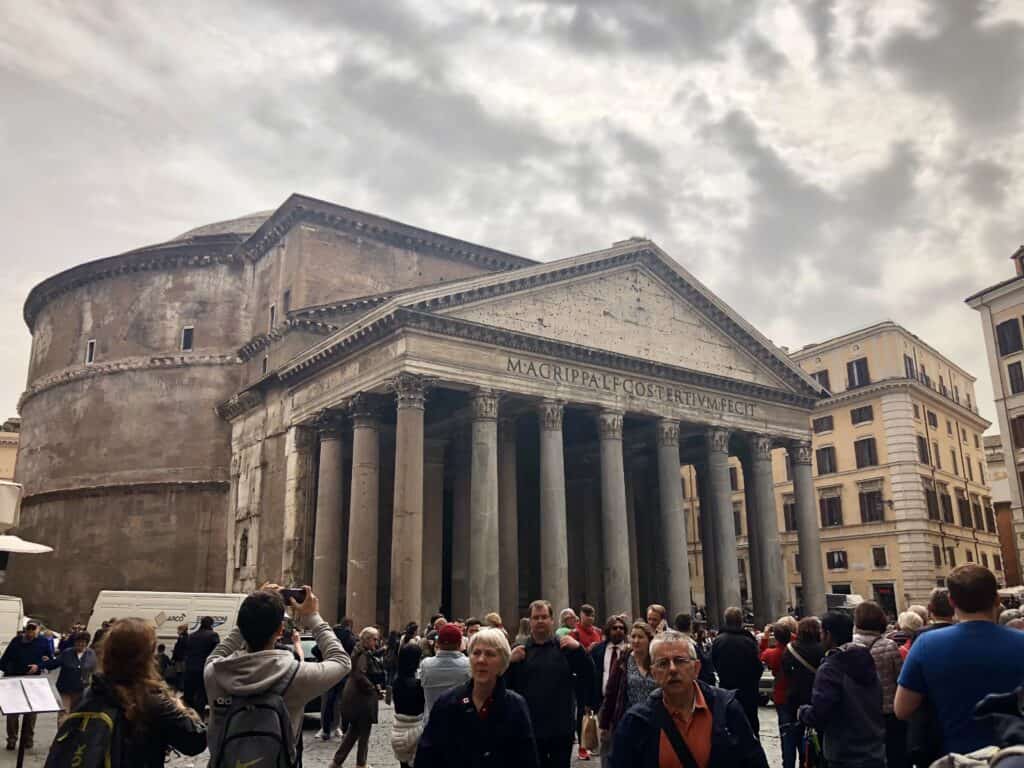 Crowds gather outside the ancient Pantheon in Rome under a cloudy sky, admiring its grand portico and iconic inscription, a must-see stop during one day in Rome. The historic structure contrasts with surrounding buildings, emphasizing its timeless architecture.