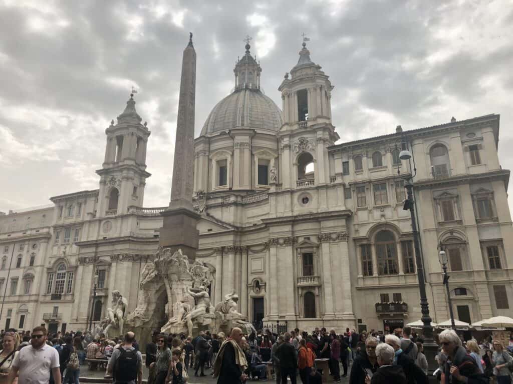 Crowds gather in Piazza Navona in front of the ornate Fountain of the Four Rivers and Sant’Agnese in Agone church, under a cloudy sky during one day in Rome. The tall obelisk and dramatic sculptures make this a vibrant and artistic stop in the historic city center.