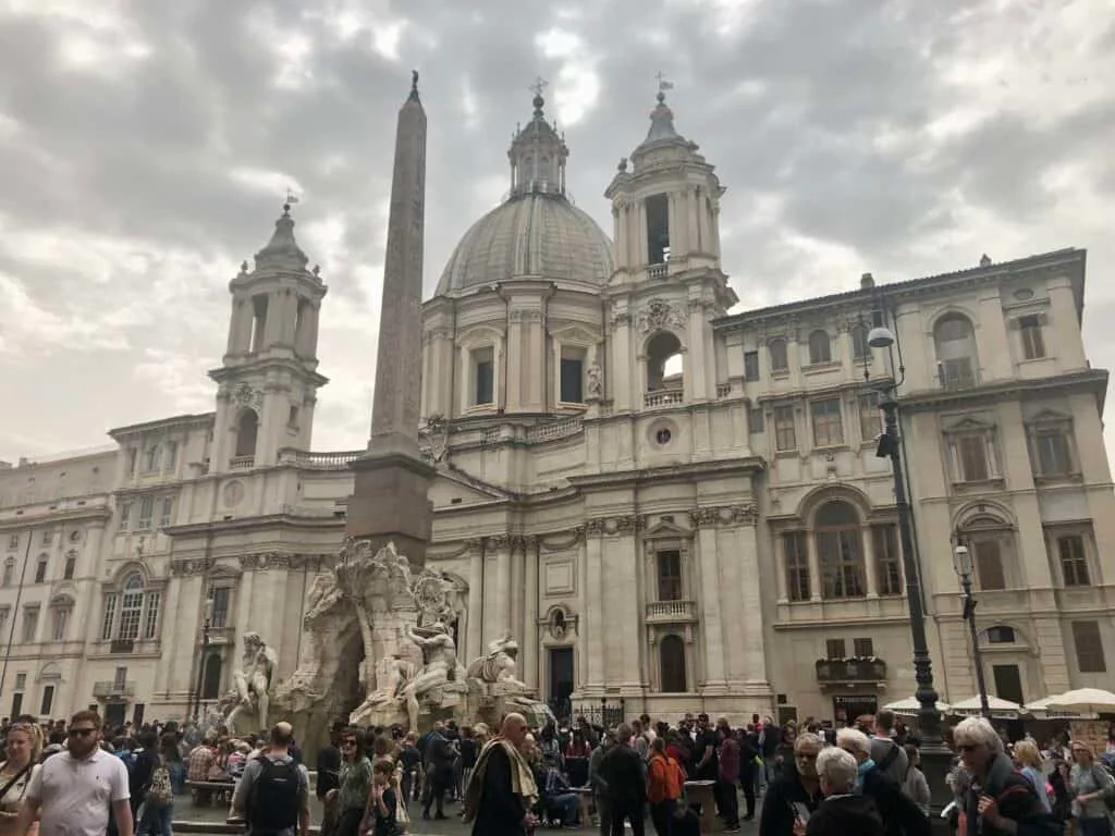 Crowds gather in Piazza Navona in front of the ornate Fountain of the Four Rivers and Sant’Agnese in Agone church, under a cloudy sky during one day in Rome. The tall obelisk and dramatic sculptures make this a vibrant and artistic stop in the historic city center.