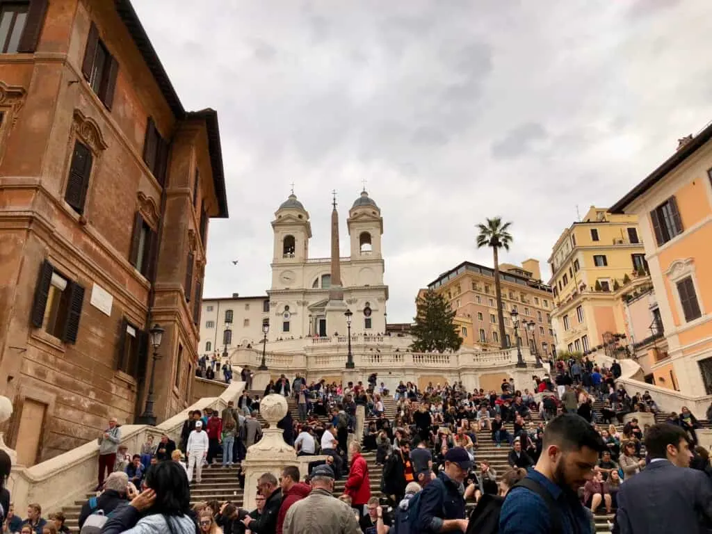 A large crowd sits and strolls along the Spanish Steps in Rome, leading up to the Trinità dei Monti church with its twin bell towers, a lively and iconic scene during one day in Rome. The surrounding pastel-colored buildings and palm tree add charm to the historic piazza.