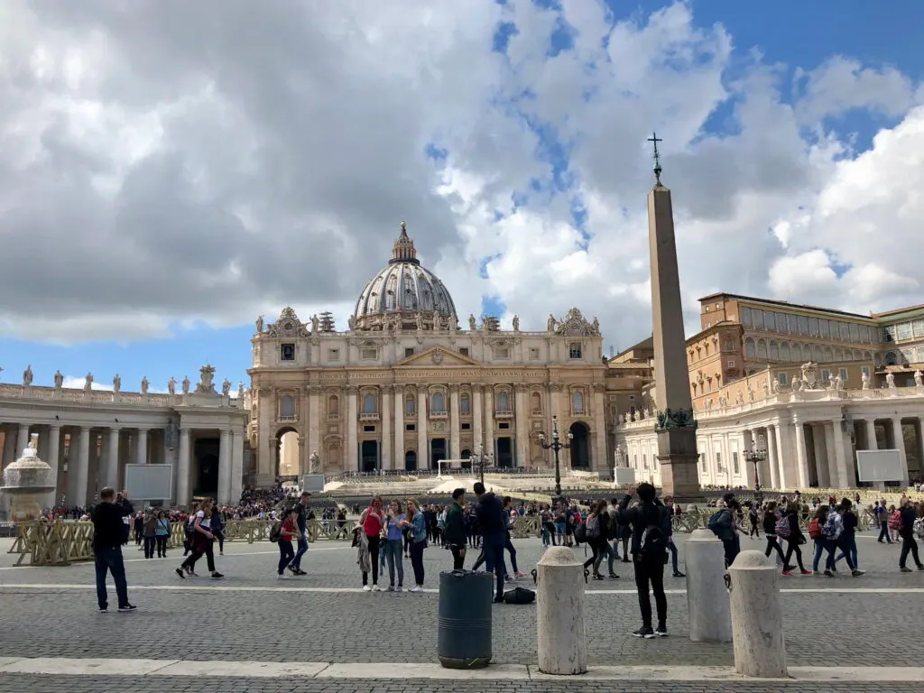 Tourists walking through St. Peter's Square with St. Peter's Basilica and the Vatican Obelisk in view under a partly cloudy sky, capturing a popular stop during one day in Rome. The scene is lively with people taking photos and admiring the grand architecture.