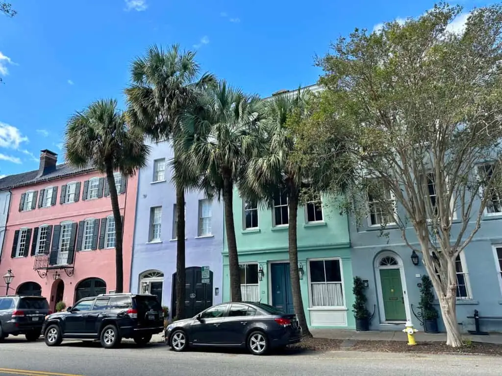 Colorful pastel buildings and palm trees line Rainbow Row in Charleston, South Carolina, with parked cars along the quiet street under a clear blue sky. Charleston’s historic charm and haunted legends make it a standout among the best Halloween towns in the USA.