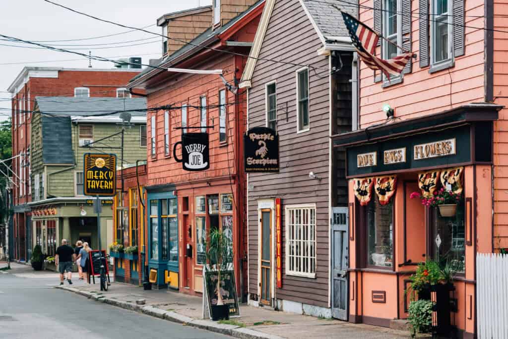A row of colorful historic buildings in Salem, Massachusetts, featuring eclectic shops like Mercy Tavern, Derby Joe, and Dave Binks Flowers, with festive bunting and flower boxes lining the sidewalk. This charming downtown scene reflects the cozy, spooky vibe that makes Salem one of the best Halloween towns in the USA.