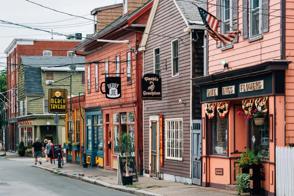 A row of colorful historic buildings in Salem, Massachusetts, featuring eclectic shops like Mercy Tavern, Derby Joe, and Dave Binks Flowers, with festive bunting and flower boxes lining the sidewalk. This charming downtown scene reflects the cozy, spooky vibe that makes Salem one of the best Halloween towns in the USA.
