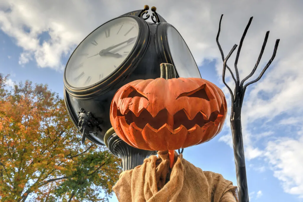 Close-up of a spooky jack-o'-lantern scarecrow with a jagged grin, set in front of a vintage street clock under a cloudy autumn sky. This festive Halloween decoration captures the spirited charm found in the best Halloween towns in the USA.